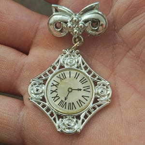 May include: Silver-toned vintage brooch featuring a clock design. The brooch has a bow-shaped top and a diamond-shaped frame around the clock face. The clock face displays Roman numerals and hands pointing to approximately 3:15.