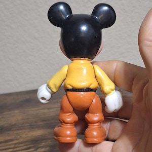 Puede incluir: Una figura vintage de Mickey Mouse, vista desde atr&aacute;s. La figura tiene orejas y cabeza negras, una camisa amarilla, pantalones naranjas y guantes blancos. Un juguete coleccionable cl&aacute;sico de Disney.