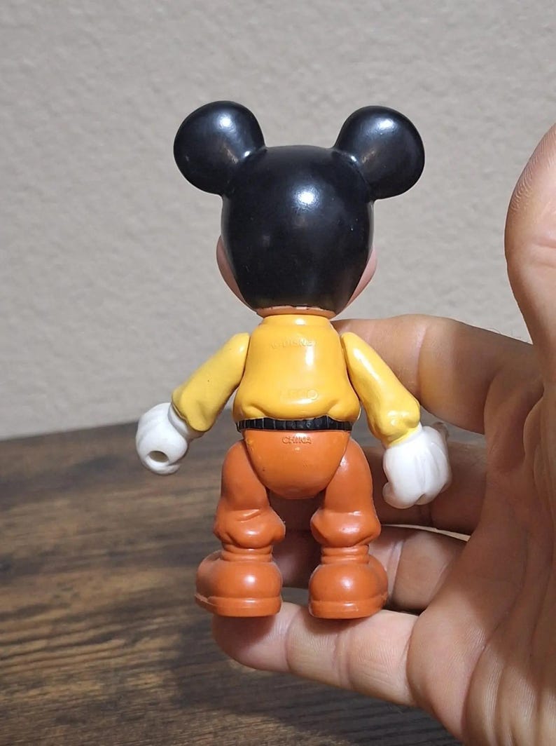 Puede incluir: Una figura vintage de Mickey Mouse, vista desde atr&aacute;s. La figura tiene orejas y cabeza negras, una camisa amarilla, pantalones naranjas y guantes blancos. Un juguete coleccionable cl&aacute;sico de Disney.