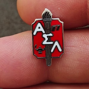 May include: A small rectangular metal lapel pin with a red enamel background featuring a vertical torch graphic. The pin displays the white Greek letters Alpha, Sigma, and Lambda, along with a small black lamp icon positioned on the lower left side.