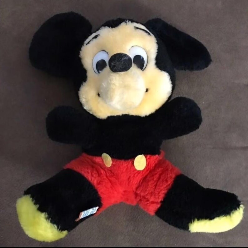 May include: A plush Mickey Mouse toy with black ears, a yellow face, and red shorts. The toy has large, black eyes and a black nose.