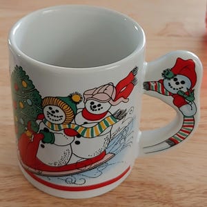 May include: White ceramic mug with a festive winter design. The mug features a snowman theme with snowmen sledding, a Christmas tree, and a snowman on the handle. The design includes red, green, and yellow accents, perfect for holiday beverages.