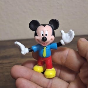 May include: A small Mickey Mouse figurine. The figure has black ears, a blue shirt with a yellow tie, red pants, yellow shoes, and white gloves. The figure is in a standing pose with arms outstretched.