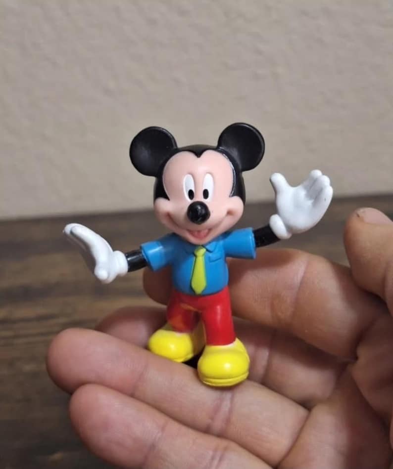 May include: A small Mickey Mouse figurine. The figure has black ears, a blue shirt with a yellow tie, red pants, yellow shoes, and white gloves. The figure is in a standing pose with arms outstretched.