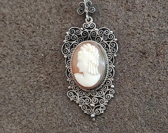 May include: A silver pendant featuring a cameo of a woman's profile. The cameo is set in an oval frame with a detailed, swirling filigree design. The pendant has a decorative bail at the top.