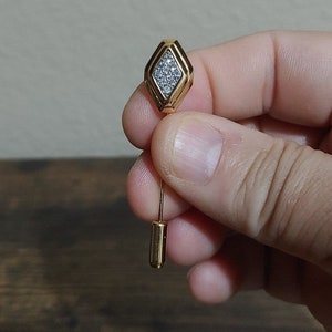 May include: A gold-toned pin with a diamond-shaped head set with clear rhinestones. The pin has a long, thin stem.