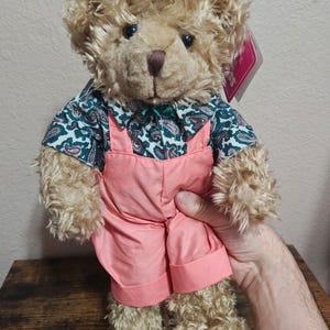 May include: A plush teddy bear with light brown fur, wearing a short-sleeved shirt featuring a green and white paisley pattern and solid pink overalls with cuffed hems. A small pink tag is attached to the bear's ear area, partially visible.