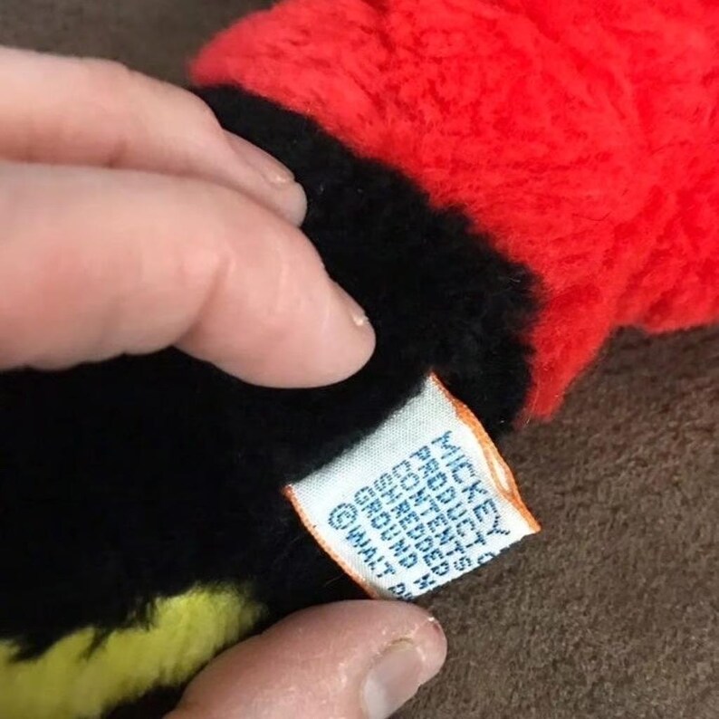 May include: Close-up of a black, red, and yellow plush toy with a tag that reads "Mickey Mouse Products, Contents: Shredded Ground Peanuts, Walt Disney Productions".