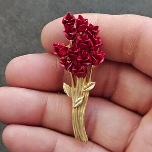 May include: A gold-toned brooch featuring a bouquet of red roses. The roses are clustered together, with detailed petals and a glossy finish. The stems and leaves are also gold, creating a striking contrast.
