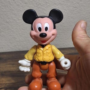 Puede incluir: Una figura de acci&oacute;n de Mickey Mouse con una camisa amarilla, pantalones marrones y orejas negras. La figura tiene guantes blancos y zapatos naranjas. El juguete se sostiene en una mano.