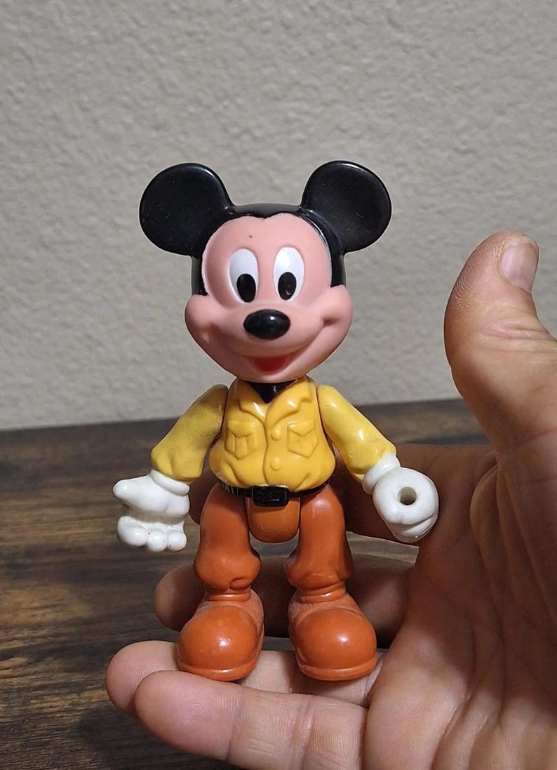 Puede incluir: Una figura de acci&oacute;n de Mickey Mouse con una camisa amarilla, pantalones marrones y orejas negras. La figura tiene guantes blancos y zapatos naranjas. El juguete se sostiene en una mano.