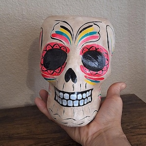 May include: A white ceramic skull with a colorful Day of the Dead design. The skull has black eyes, red and pink floral patterns, and yellow and blue accents.