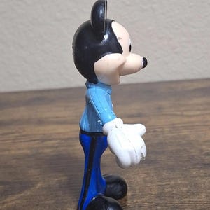 Puede incluir: Una peque&ntilde;a figura de Mickey Mouse. La figura tiene cabeza y orejas negras, cara blanca y nariz negra. Viste chaqueta azul, pantalones azules, guantes blancos y zapatos negros. La figura est&aacute; sobre una superficie de madera.