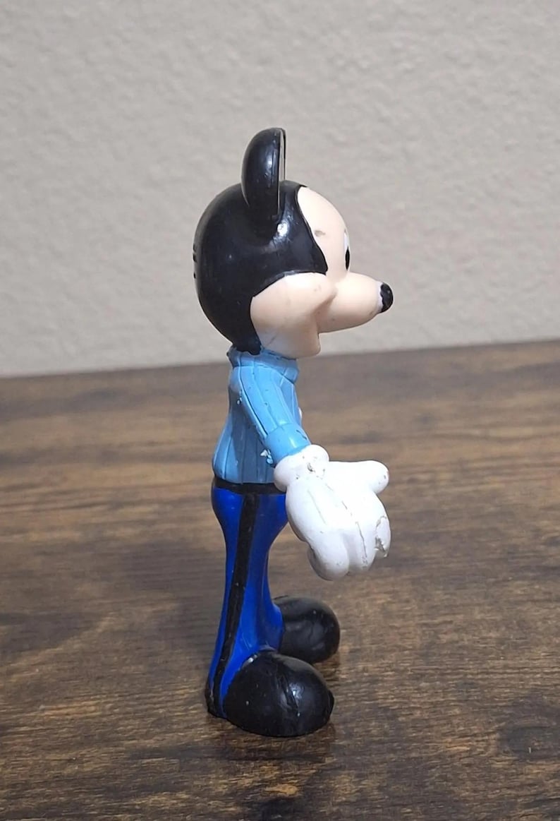 Puede incluir: Una peque&ntilde;a figura de Mickey Mouse. La figura tiene cabeza y orejas negras, cara blanca y nariz negra. Viste chaqueta azul, pantalones azules, guantes blancos y zapatos negros. La figura est&aacute; sobre una superficie de madera.