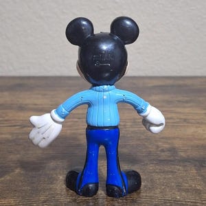 Puede incluir: Una figura de Mickey Mouse, vista desde atr&aacute;s, con orejas y cabeza negras, una parte superior azul claro, pantalones azules, guantes blancos y zapatos negros. La figura est&aacute; sobre una superficie de madera. El texto "WALT DISNEY PRODUCTIONS" es visible en la parte posterior de la cabeza.