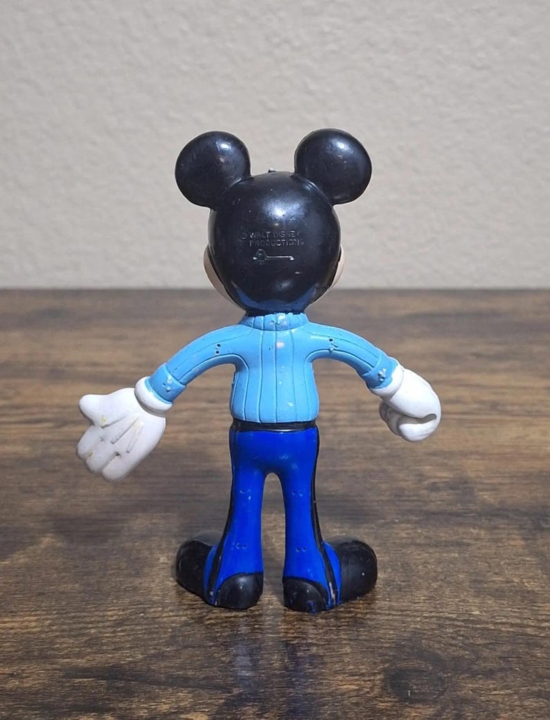 Puede incluir: Una figura de Mickey Mouse, vista desde atr&aacute;s, con orejas y cabeza negras, una parte superior azul claro, pantalones azules, guantes blancos y zapatos negros. La figura est&aacute; sobre una superficie de madera. El texto "WALT DISNEY PRODUCTIONS" es visible en la parte posterior de la cabeza.