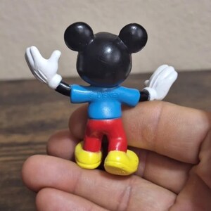 May include: A small Mickey Mouse figurine, viewed from the back, with arms outstretched. The figure has a black head with large ears, a blue shirt, red pants, yellow shoes, and white gloves. The word "DISNEY" is printed on the back of the shirt.