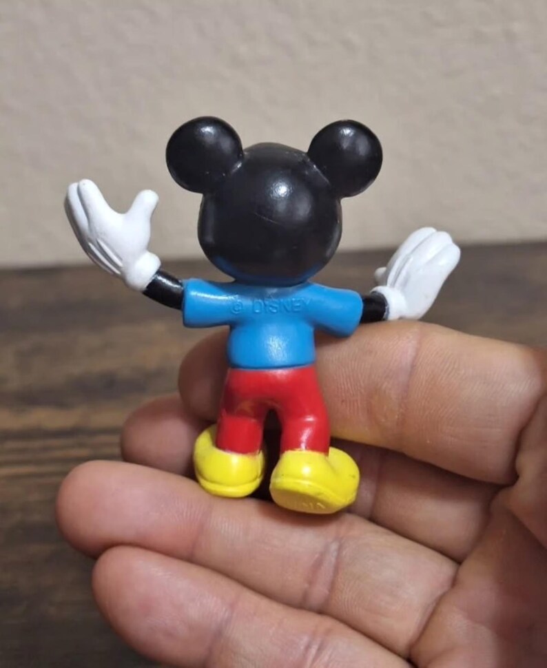 May include: A small Mickey Mouse figurine, viewed from the back, with arms outstretched. The figure has a black head with large ears, a blue shirt, red pants, yellow shoes, and white gloves. The word "DISNEY" is printed on the back of the shirt.