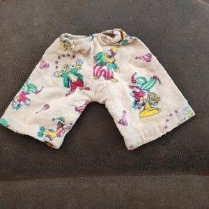 May include: Cream-colored shorts featuring a repeating pattern of colorful clowns in various poses, including juggling and playing instruments. The fabric has a textured, terry-cloth-like appearance with an elasticized waistband at the top.