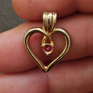 May include: A gold-colored heart-shaped pendant with a small, round, red gemstone at its center. The pendant has a loop at the top for a chain. The heart shape is open, with a twisted design at the top.