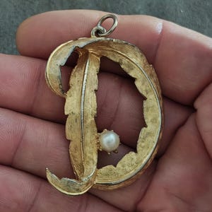 May include: Gold-tone pendant in the shape of the letter "D", with a textured, leaf-like design. A small, round, white pearl is set in the center. The pendant has a loop at the top for a chain. The overall design is vintage and elegant.