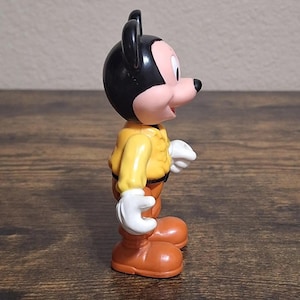 Puede incluir: Una figura vintage de Mickey Mouse. La figura tiene una cabeza negra con orejas grandes, una cara rosada y una nariz negra. Viste una chaqueta amarilla, pantalones marrones, zapatos naranjas y guantes blancos. La figura est&aacute; sobre una superficie de madera.