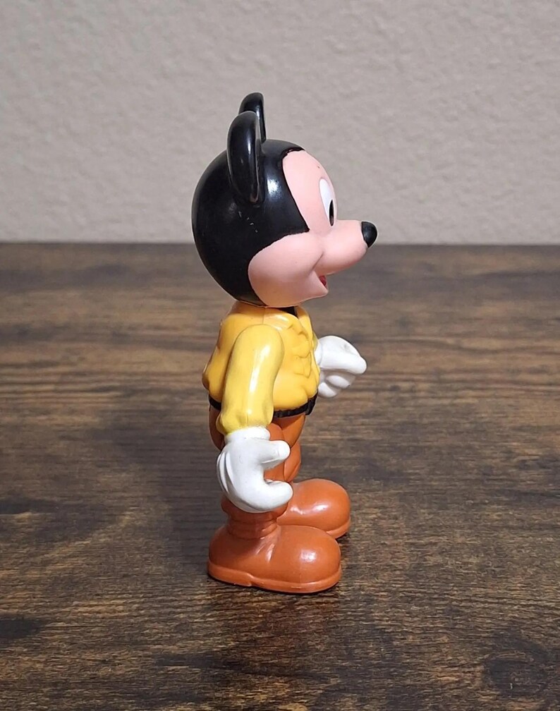 Puede incluir: Una figura vintage de Mickey Mouse. La figura tiene una cabeza negra con orejas grandes, una cara rosada y una nariz negra. Viste una chaqueta amarilla, pantalones marrones, zapatos naranjas y guantes blancos. La figura est&aacute; sobre una superficie de madera.