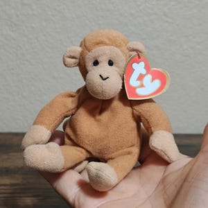 May include: A small, plush brown monkey toy with a heart-shaped tag. The monkey has a tan face, ears, hands, and feet. It is sitting with its arms and legs outstretched. The tag is red and white with the letters "Ty".