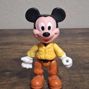 Puede incluir: Una figura de Mickey Mouse con camisa amarilla, pantalones marrones y orejas negras. La figura tiene guantes blancos y zapatos naranjas. El juguete est&aacute; sobre una superficie de madera.