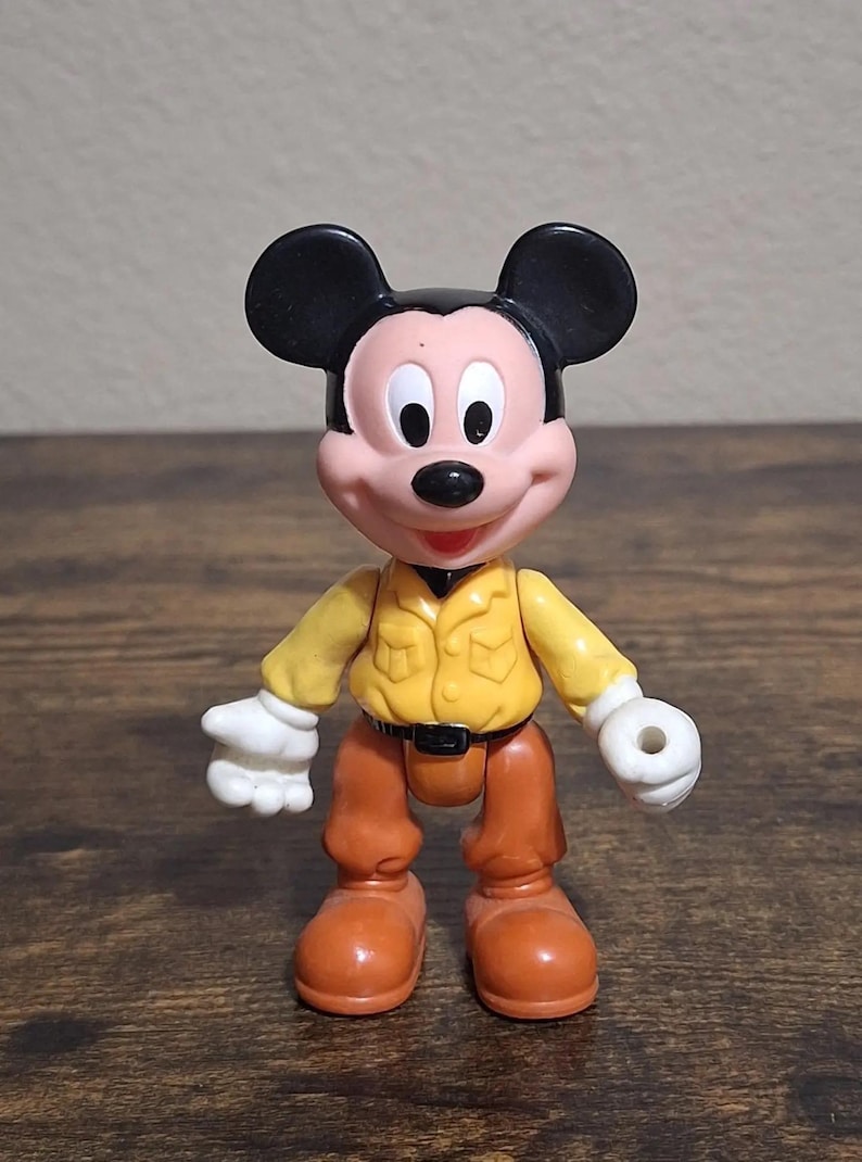 Puede incluir: Una figura de Mickey Mouse con camisa amarilla, pantalones marrones y orejas negras. La figura tiene guantes blancos y zapatos naranjas. El juguete est&aacute; sobre una superficie de madera.
