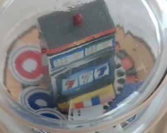 May include: A clear glass jar containing miniature casino-themed items. A small slot machine with the number 7 is visible, along with playing cards, poker chips, and other game-related objects. The jar has a small red dot on top.