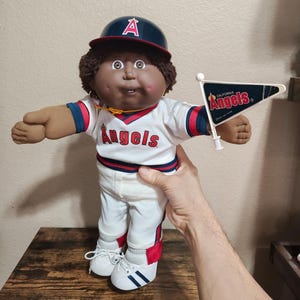 May include: A vintage Cabbage Patch Kids doll wearing a baseball uniform. The doll has brown skin, brown hair, and is wearing a navy blue baseball cap with a red "A". The uniform is white with red and blue accents, and the word "Angels" is across the chest.