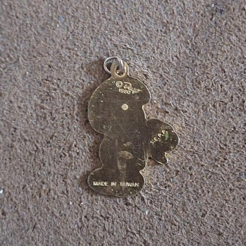 May include: A vintage gold-colored metal charm in the shape of a cartoon character. The charm has a loop at the top for attachment and the text "&copy; OZ 1980 S.E.P.C." and "MADE IN TAIWAN" are visible.