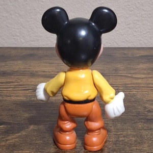 Puede incluir: Figura de Mickey Mouse vista desde atr&aacute;s. La figura tiene una cabeza negra con orejas grandes, una camisa amarilla, pantalones naranjas, guantes blancos y zapatos marrones. El juguete est&aacute; sobre una superficie de madera.
