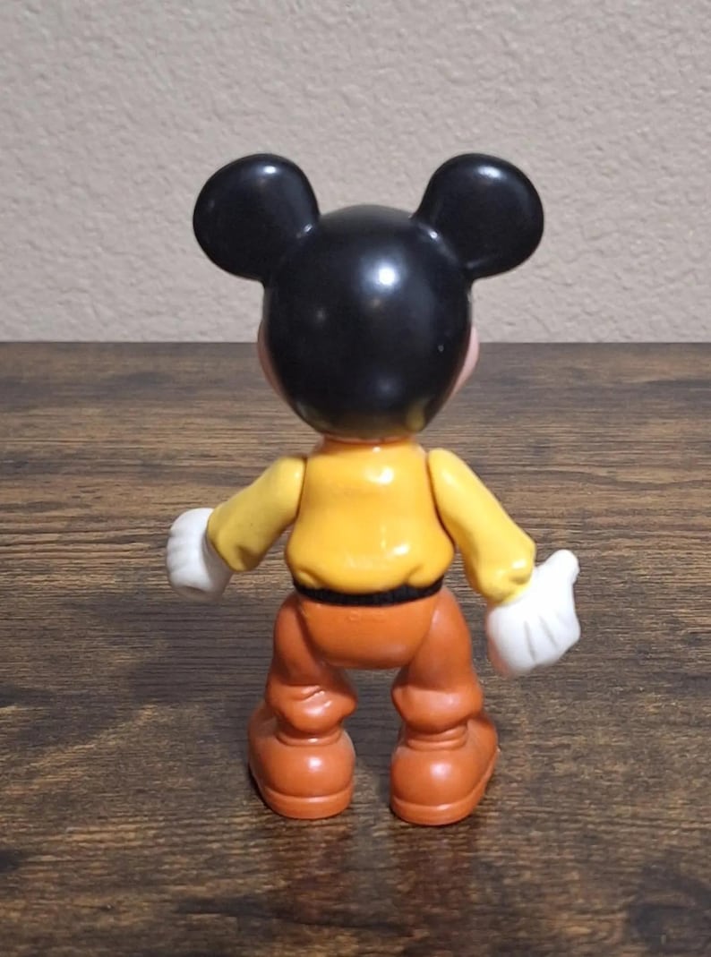 Puede incluir: Figura de Mickey Mouse vista desde atr&aacute;s. La figura tiene una cabeza negra con orejas grandes, una camisa amarilla, pantalones naranjas, guantes blancos y zapatos marrones. El juguete est&aacute; sobre una superficie de madera.