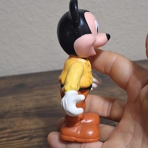 Puede incluir: Una figura vintage de Mickey Mouse. La figura tiene una cabeza negra con orejas grandes, una cara rosada y una chaqueta amarilla. Lleva pantalones y zapatos naranjas, y guantes blancos. La figura se sostiene en una mano.