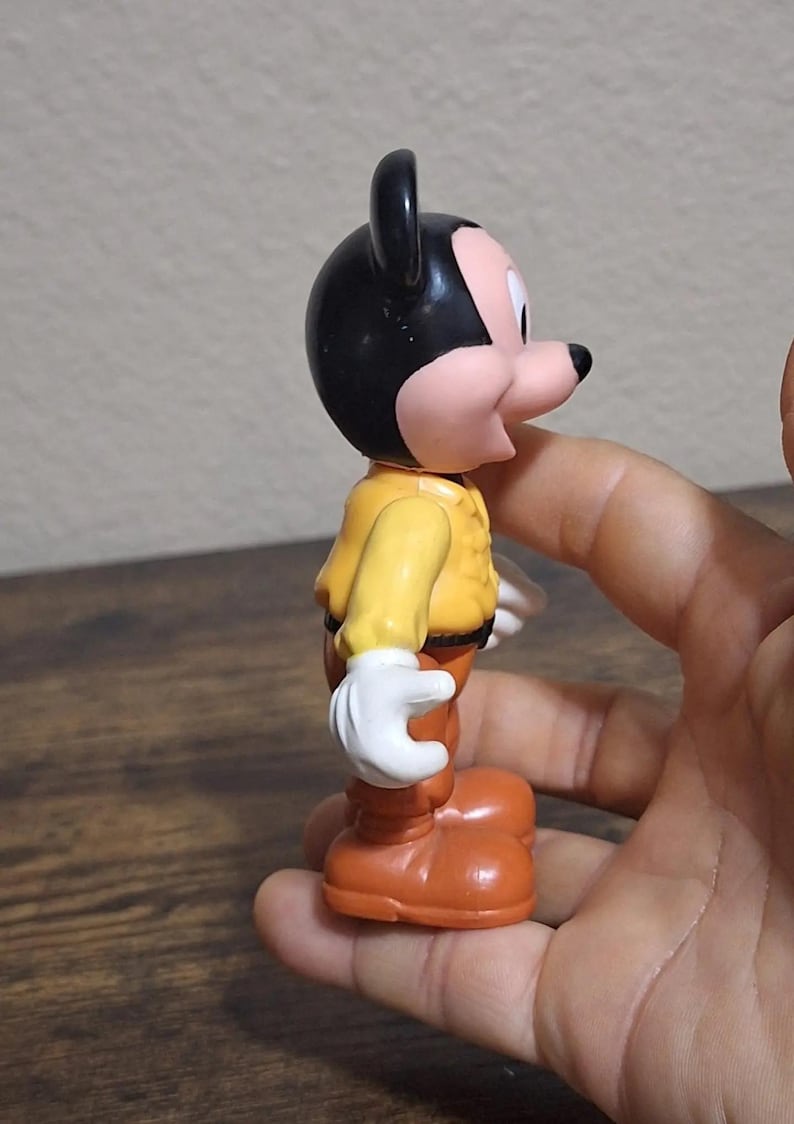 Puede incluir: Una figura vintage de Mickey Mouse. La figura tiene una cabeza negra con orejas grandes, una cara rosada y una chaqueta amarilla. Lleva pantalones y zapatos naranjas, y guantes blancos. La figura se sostiene en una mano.
