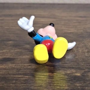 May include: A small, colorful Mickey Mouse figurine lying on its back. The figure has a pink face, black ears, and a blue jacket. It wears red shorts, yellow shoes, and white gloves. The figurine is on a brown wooden surface.