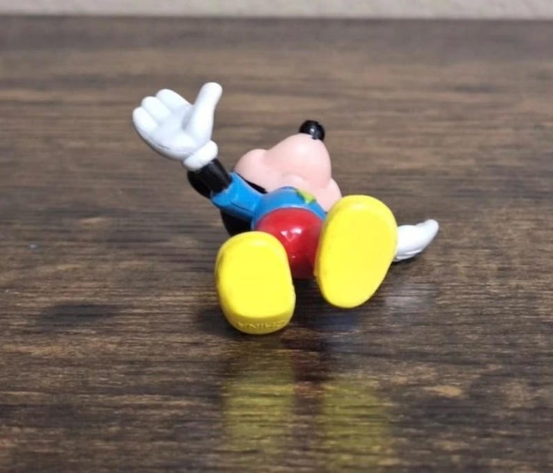 May include: A small, colorful Mickey Mouse figurine lying on its back. The figure has a pink face, black ears, and a blue jacket. It wears red shorts, yellow shoes, and white gloves. The figurine is on a brown wooden surface.