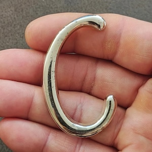 May include: A shiny, silver-colored metal letter "C" is held in a person's hand. The letter has a rounded, tubular design with a smooth, reflective surface. The "C" shape is slightly curved and has a polished appearance.