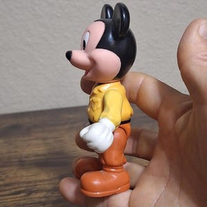 Puede incluir: Una figura vintage de Mickey Mouse. La figura tiene cabeza y orejas negras, cara rosa y chaqueta amarilla. Lleva pantalones y botas naranjas, y guantes blancos. La figura se sostiene en una mano.