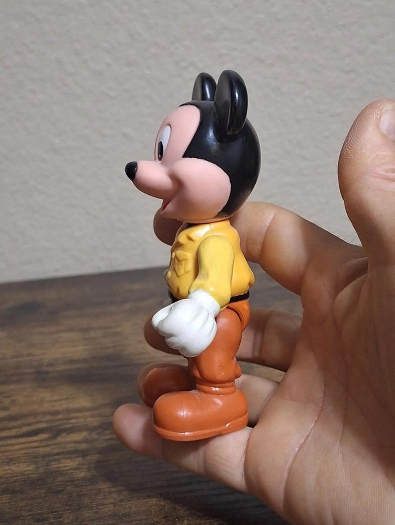 Puede incluir: Una figura vintage de Mickey Mouse. La figura tiene cabeza y orejas negras, cara rosa y chaqueta amarilla. Lleva pantalones y botas naranjas, y guantes blancos. La figura se sostiene en una mano.