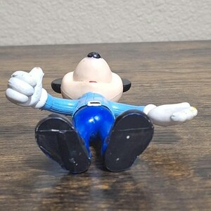 Puede incluir: Una figura de un personaje de dibujos animados tumbado boca arriba con los brazos extendidos. La figura tiene una cara bronceada, orejas negras y viste un traje azul con zapatos negros y guantes blancos. La figura est&aacute; sobre una superficie de madera.