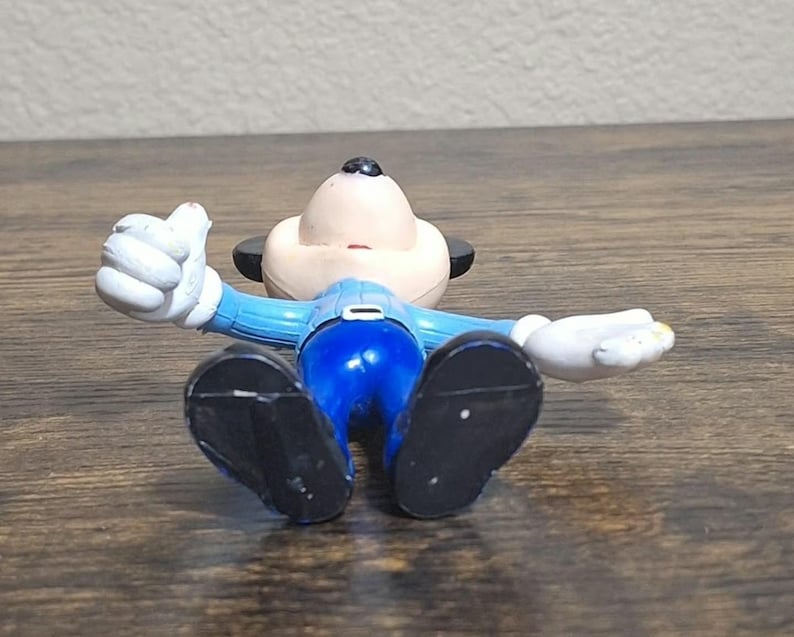 Puede incluir: Una figura de un personaje de dibujos animados tumbado boca arriba con los brazos extendidos. La figura tiene una cara bronceada, orejas negras y viste un traje azul con zapatos negros y guantes blancos. La figura est&aacute; sobre una superficie de madera.