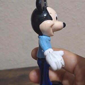 Puede incluir: Una figura vintage de Mickey Mouse. La figura lleva un su&eacute;ter azul claro, pantalones azul oscuro y zapatos negros. La figura tiene orejas negras y una cara y guantes blancos. La figura se sostiene en una mano.