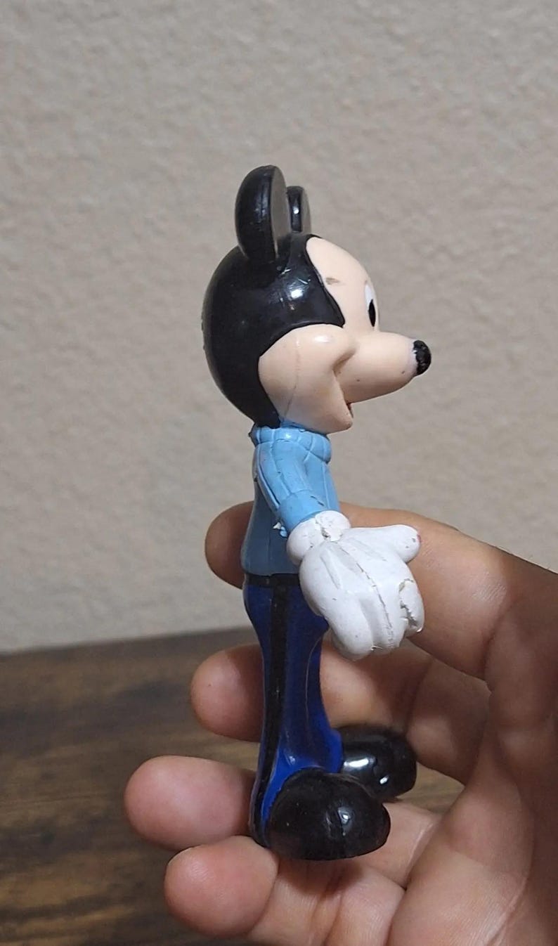 Puede incluir: Una figura vintage de Mickey Mouse. La figura lleva un su&eacute;ter azul claro, pantalones azul oscuro y zapatos negros. La figura tiene orejas negras y una cara y guantes blancos. La figura se sostiene en una mano.