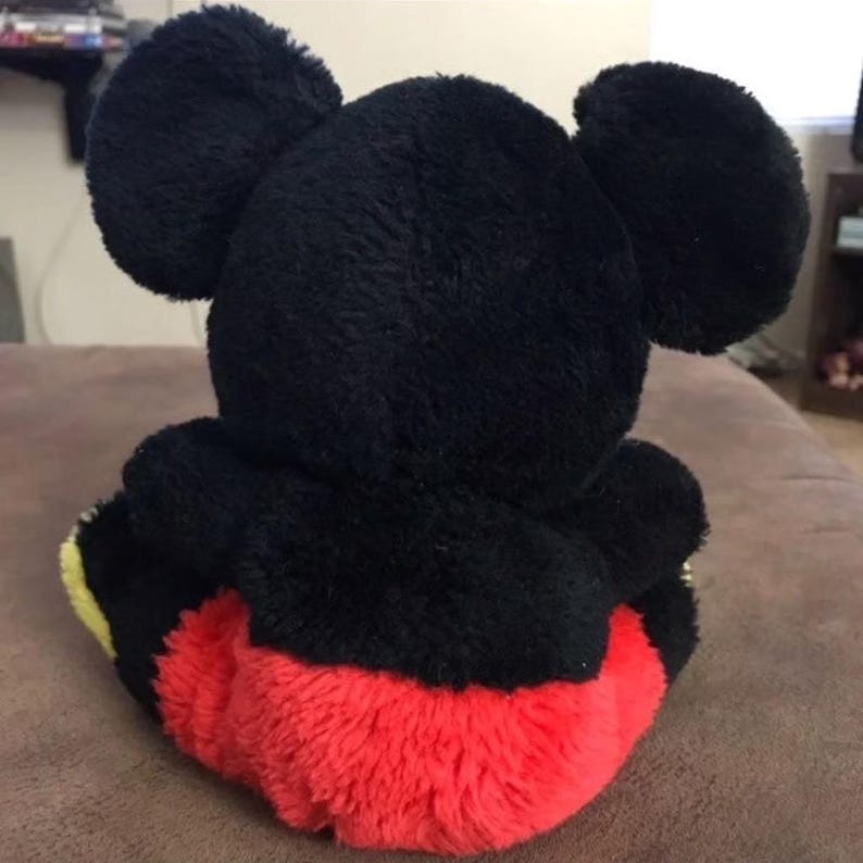 May include: A black and red plush Mickey Mouse toy. The toy is sitting on a brown surface. The toy has large black ears and a red and yellow body.