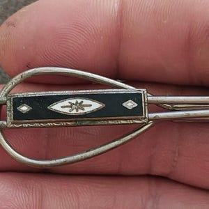 May include: A silver-toned metal tie clip featuring an elongated teardrop-shaped frame. The center bar is inlaid with a rectangular black panel, decorated with a white eye-shaped motif containing a central starburst and two small diamond accents.