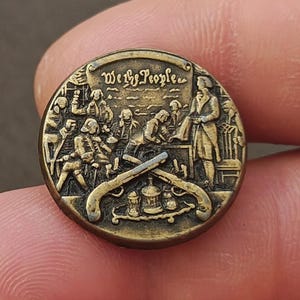 May include: A round, antique brass button with a detailed relief scene. The button features the text "We the People" above a depiction of figures gathered around a table, with crossed pistols and a lamp. The button has a vintage, aged appearance.