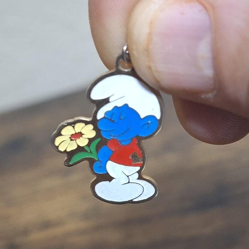 May include: A Smurf charm pendant featuring a blue Smurf holding a yellow flower. The Smurf is wearing a red shirt, white pants, and a white hat. The pendant has a silver-colored metal finish and a small loop for attaching to a chain or bracelet.